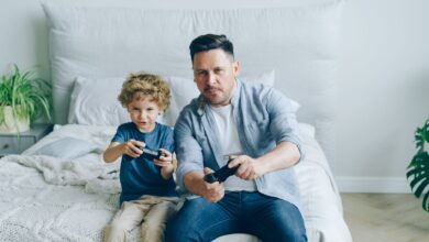 Father and son joyfully playing video games while sitting on a bed, capturing a moment of family fun.
