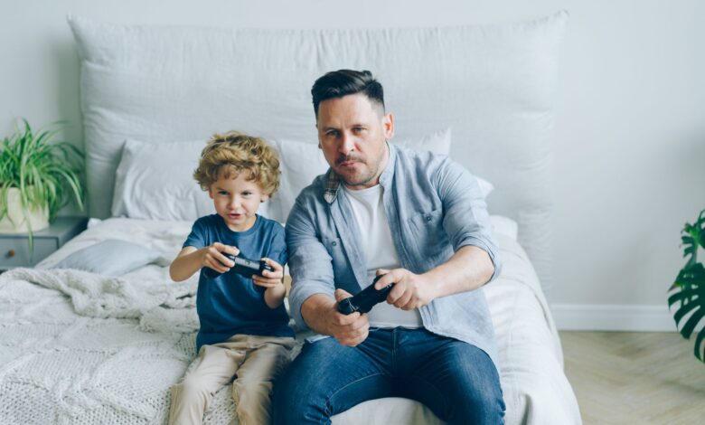 Father and son joyfully playing video games while sitting on a bed, capturing a moment of family fun.