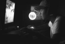Side view of a gamer wearing a cap and headphones, illuminated by a computer screen in a dark room.