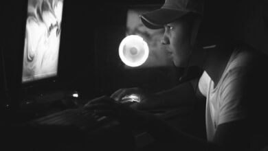 Side view of a gamer wearing a cap and headphones, illuminated by a computer screen in a dark room.