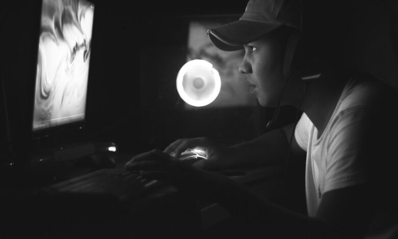 Side view of a gamer wearing a cap and headphones, illuminated by a computer screen in a dark room.