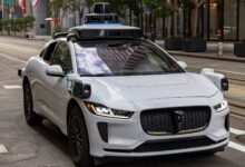 A white autonomous vehicle navigating a city street, reflecting urban architecture in daylight.