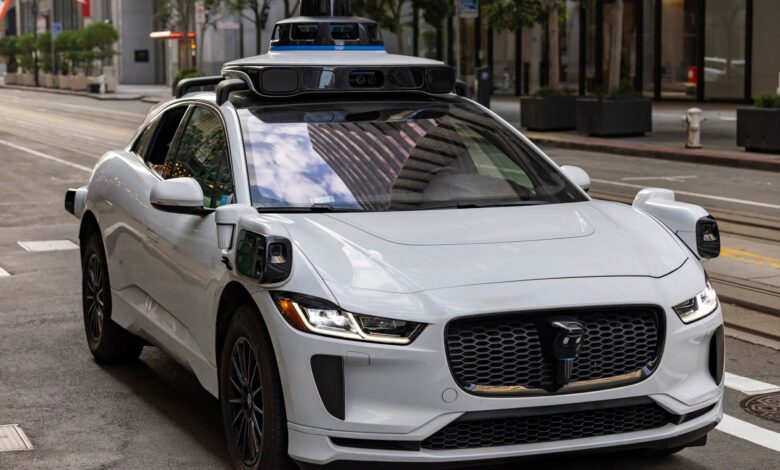 A white autonomous vehicle navigating a city street, reflecting urban architecture in daylight.