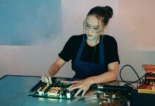 Young woman in protective goggles soldering electronics on a workbench indoors.