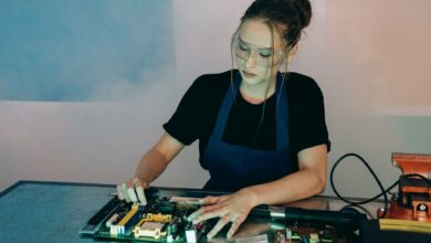 Young woman in protective goggles soldering electronics on a workbench indoors.