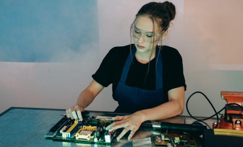 Young woman in protective goggles soldering electronics on a workbench indoors.