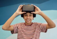 Cheerful little boy in t shirt putting pn VR glasses and looking at camera against blue wall