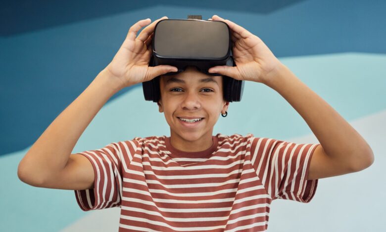 Cheerful little boy in t shirt putting pn VR glasses and looking at camera against blue wall