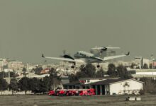 Airplane takes off over urban landscape with fire trucks ready at the fire station.