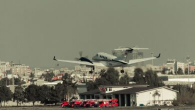 Airplane takes off over urban landscape with fire trucks ready at the fire station.