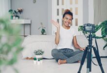 Smiling woman waves at camera while recording a vlog in a stylish bedroom setting.