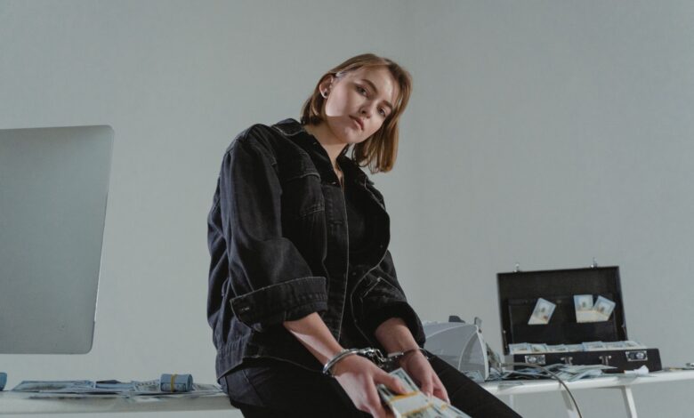 Young woman in black jacket handcuffed with cash, conceptual indoors setting.