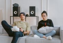 Two young adults enjoying leisure time on a sofa playing video games, surrounded by home speakers.
