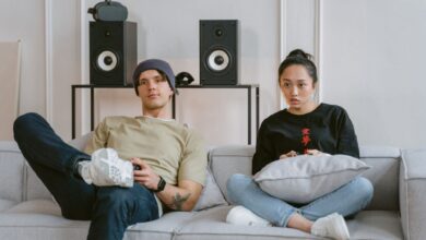 Two young adults enjoying leisure time on a sofa playing video games, surrounded by home speakers.