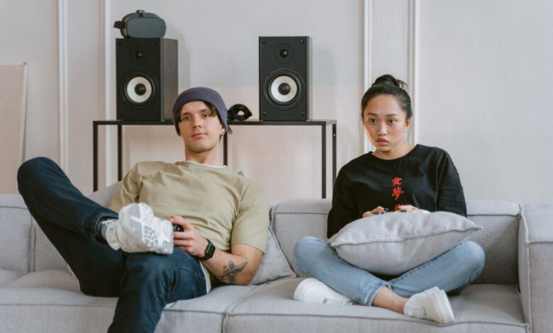 Two young adults enjoying leisure time on a sofa playing video games, surrounded by home speakers.