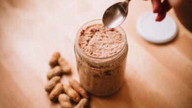 A glass jar of homemade peanut butter on a wooden table with peanuts and a spoon.