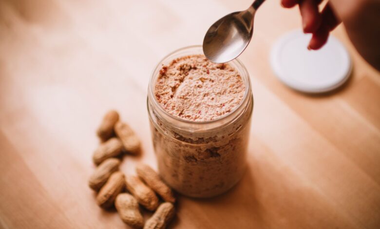 A glass jar of homemade peanut butter on a wooden table with peanuts and a spoon.