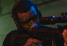 An adult man wearing eye and hearing protection focuses intently while aiming a rifle.