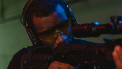 An adult man wearing eye and hearing protection focuses intently while aiming a rifle.