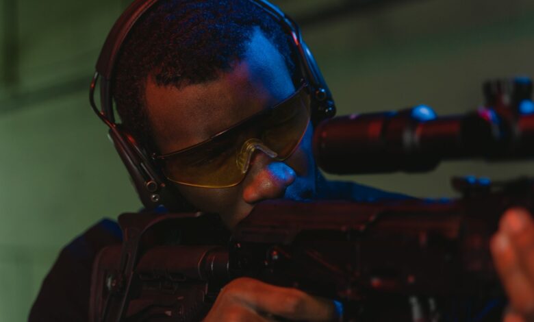 An adult man wearing eye and hearing protection focuses intently while aiming a rifle.