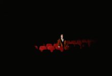 A woman in a fur coat sits alone in a dimly lit theater, surrounded by empty red seats.