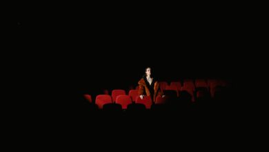 A woman in a fur coat sits alone in a dimly lit theater, surrounded by empty red seats.