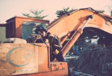 Soldier aiming a rifle outdoors, using an excavator for cover, in a tactical scene.