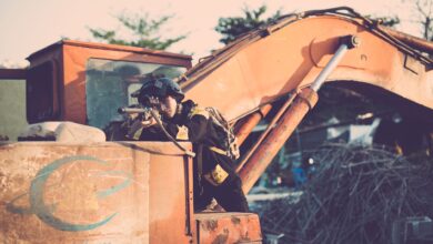 Soldier aiming a rifle outdoors, using an excavator for cover, in a tactical scene.
