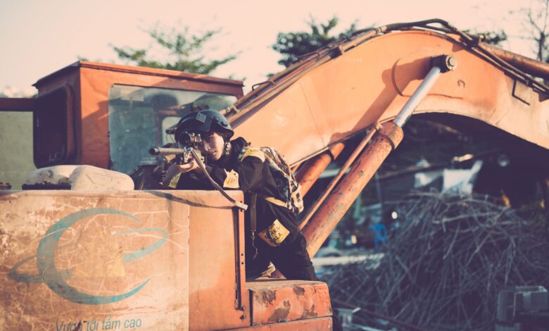 Soldier aiming a rifle outdoors, using an excavator for cover, in a tactical scene.