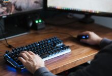 High angle crop male in casual clothes sitting at desk and browsing contemporary computer while typing on backlit keyboard and using mouse