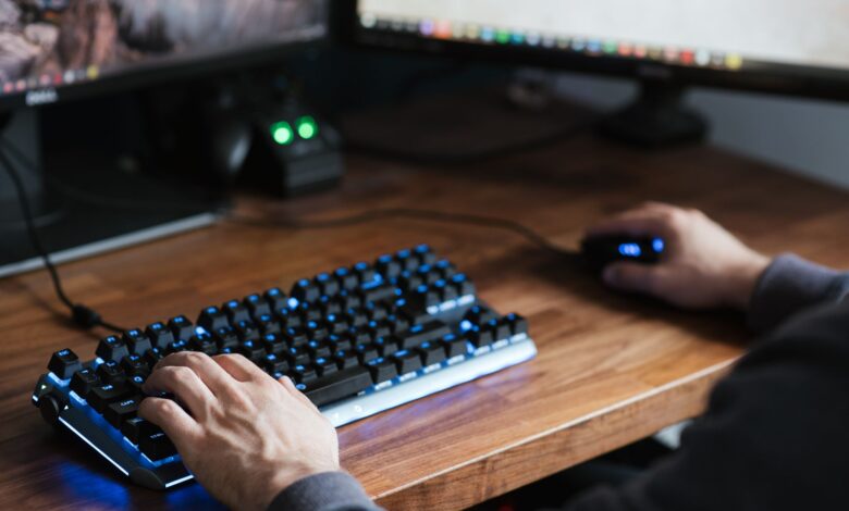High angle crop male in casual clothes sitting at desk and browsing contemporary computer while typing on backlit keyboard and using mouse
