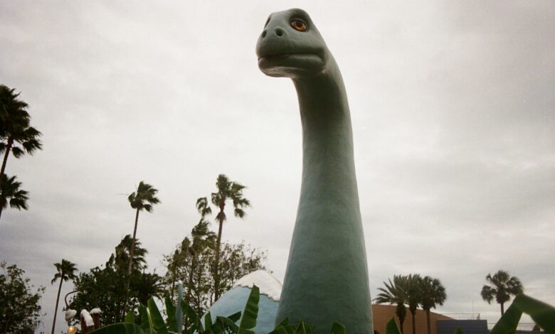 A large dinosaur statue surrounded by palm trees under a cloudy sky.
