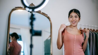 Smiling young ethnic female blogger standing in room and demonstrating pink dress while shooting vlog on smartphone with ring lamp near mirror and wardrobe