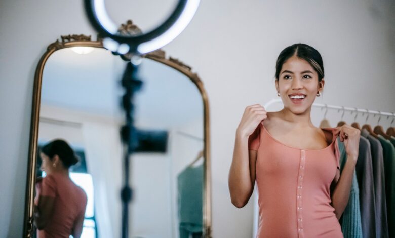 Smiling young ethnic female blogger standing in room and demonstrating pink dress while shooting vlog on smartphone with ring lamp near mirror and wardrobe