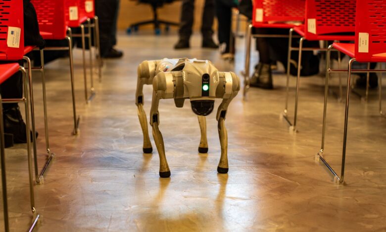 A robotic dog navigates an indoor setting amidst red chairs, showcasing technology in modern environments.