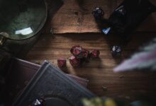 A dark, moody still life scene featuring red and blue gaming dice, books, and a magnifier on a wooden surface.