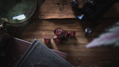 A dark, moody still life scene featuring red and blue gaming dice, books, and a magnifier on a wooden surface.