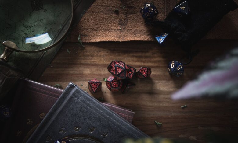 A dark, moody still life scene featuring red and blue gaming dice, books, and a magnifier on a wooden surface.