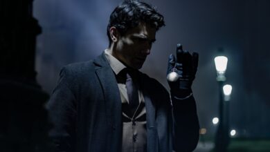 A man in dark clothing holds a pocket watch on a dimly lit street at night.