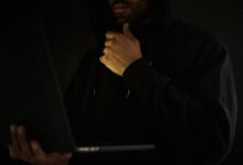 Crop contemplative male hacker in black hoodie watching netbook and touching chin thoughtfully in dark room