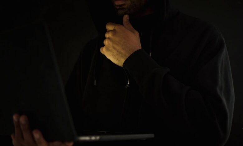 Crop contemplative male hacker in black hoodie watching netbook and touching chin thoughtfully in dark room