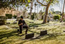 A man kneeling with a memento in a tranquil cemetery surrounded by tombstones and trees.
