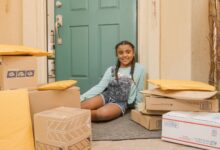 A young girl sitting on a porch surrounded by delivery packages with a cheerful expression.