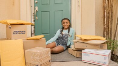 A young girl sitting on a porch surrounded by delivery packages with a cheerful expression.