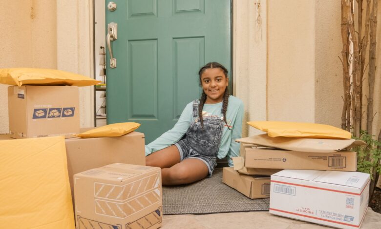 A young girl sitting on a porch surrounded by delivery packages with a cheerful expression.