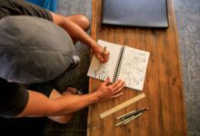A person sketches in a notebook at a wooden table, viewed from above.