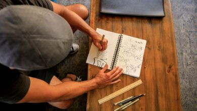 A person sketches in a notebook at a wooden table, viewed from above.