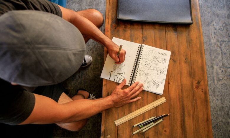 A person sketches in a notebook at a wooden table, viewed from above.