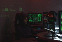 A man in a black hoodie engaged in cybersecurity work using multiple monitors indoors.