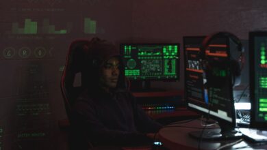 A man in a black hoodie engaged in cybersecurity work using multiple monitors indoors.